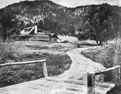 View up a dirt road to the main building of the McGraw ranch, a two story ranch-style building with an enclosed porch, rustic architecture, and a sweeping view of lumpy ridge in the background.