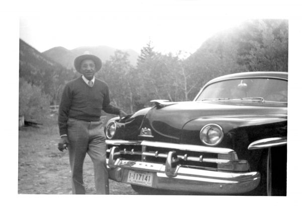 A man stands in front of a parked car in a mountain valley.