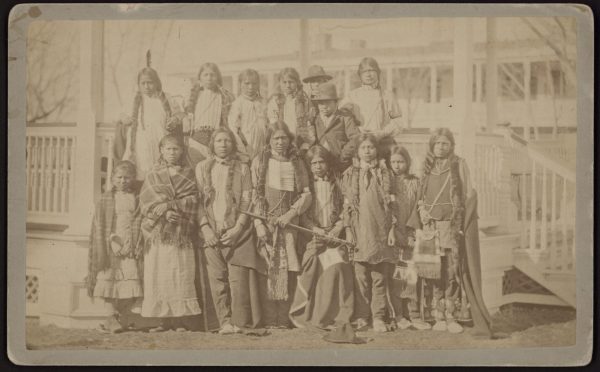 A group of Northern Arapaho and Shoshone youth on the day they were admitted to Carlisel Indian School. The students are grouped together, looking nervous but stoic. They wear traditional dress including feathers, blankets, and other Native American garb.
