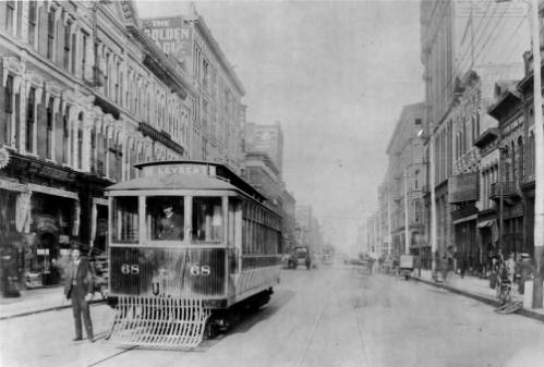 Black and white photograph of a historic trolley car on Larimer Street. The trolley runs down the left hand side of the street surrounded by two-story buildings.